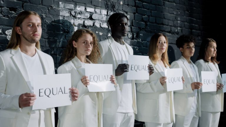 Sex unga människor i vita kläder på rad med ryggarna mot en mörk vägg. Alla håller i varsitt papper med texten "Equal"