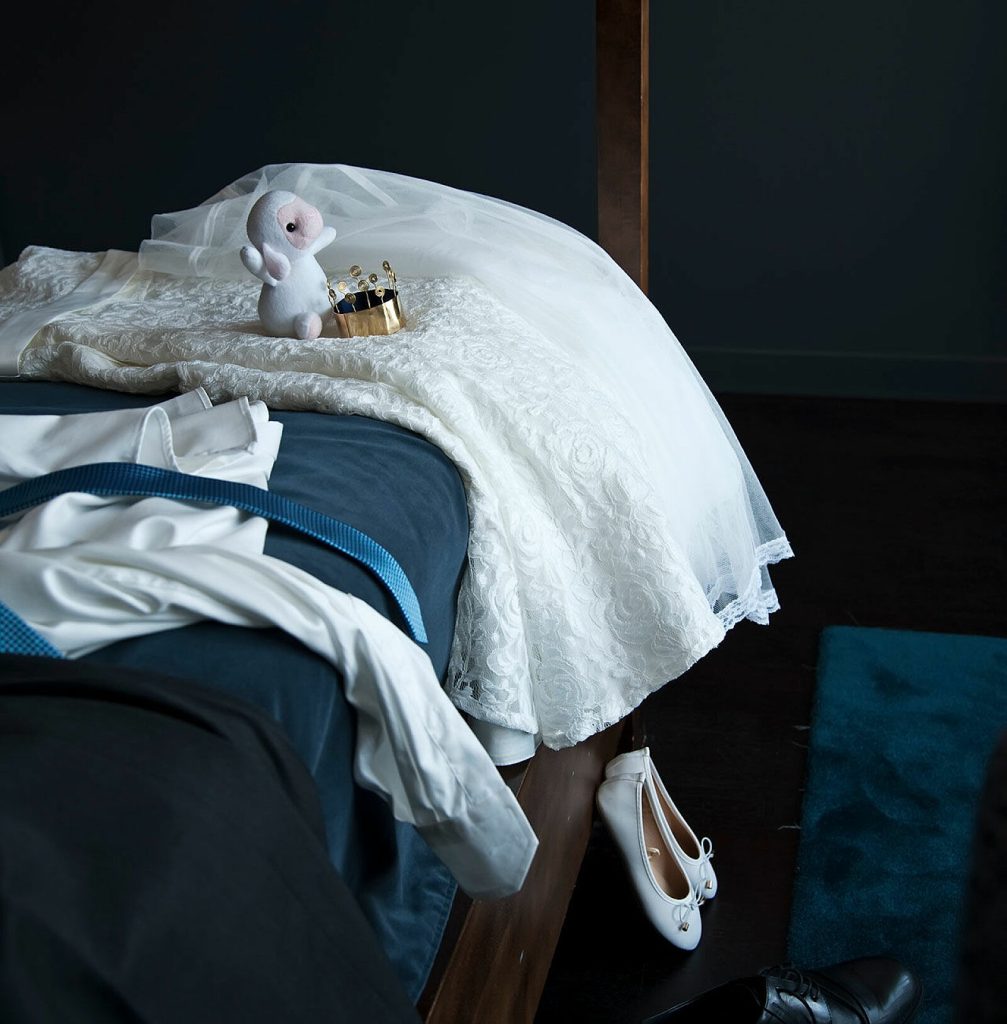Ett mörkt rum där det står en säng klädd med ett sängöverkast. Ovanpå sängen ligger en brudklänning bredvid brudgumskläder i vitt och blått. På golvet står det ett par vita finskor i barnstorlek. På brudklänningen sitter det ett vitt kramdjur intill en brudkrona.